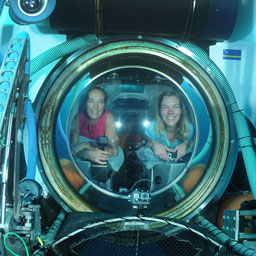 two women peer out of a porthole on a submersible underwater